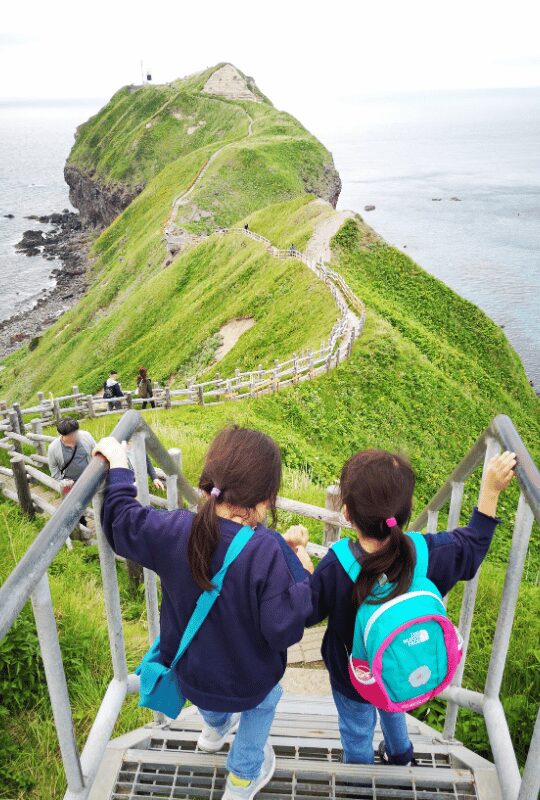 積丹岬の階段を降りる子ども2人の後ろ姿。緑の断崖と青い海が広がる