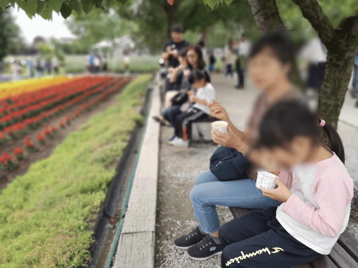 祖母と子どもが並んでソフトクリームを食べている。背後に花畑